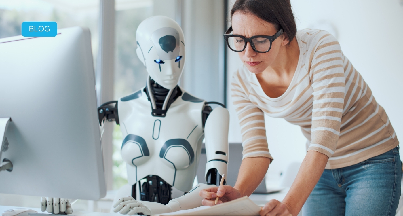 Eine Frau zeigt einem Roboter etwas auf Papier. Sie sitzen am Arbeitsplatz vor einem Computer